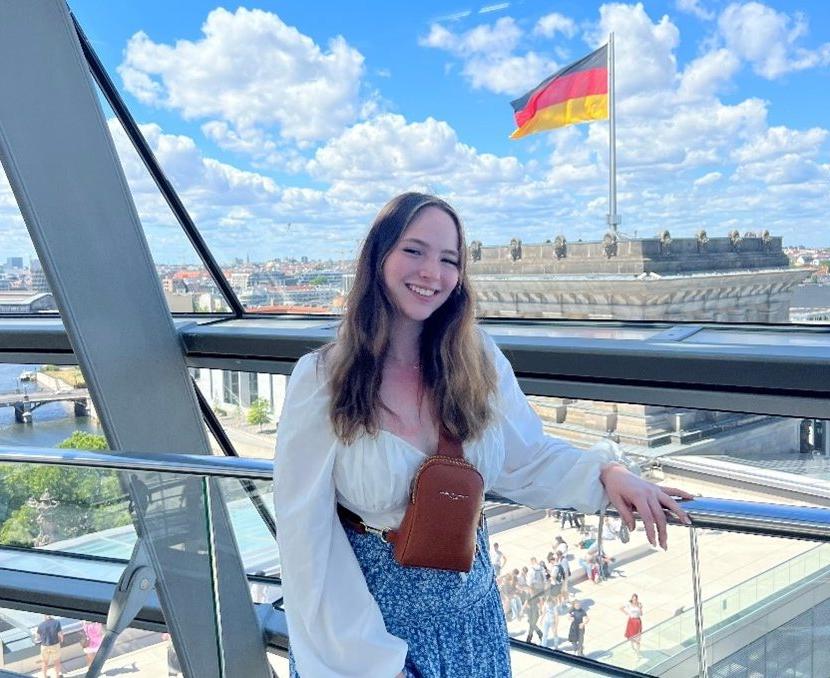 一位年轻女性站在一座玻璃桥上，她身后的建筑展示着德国国旗.